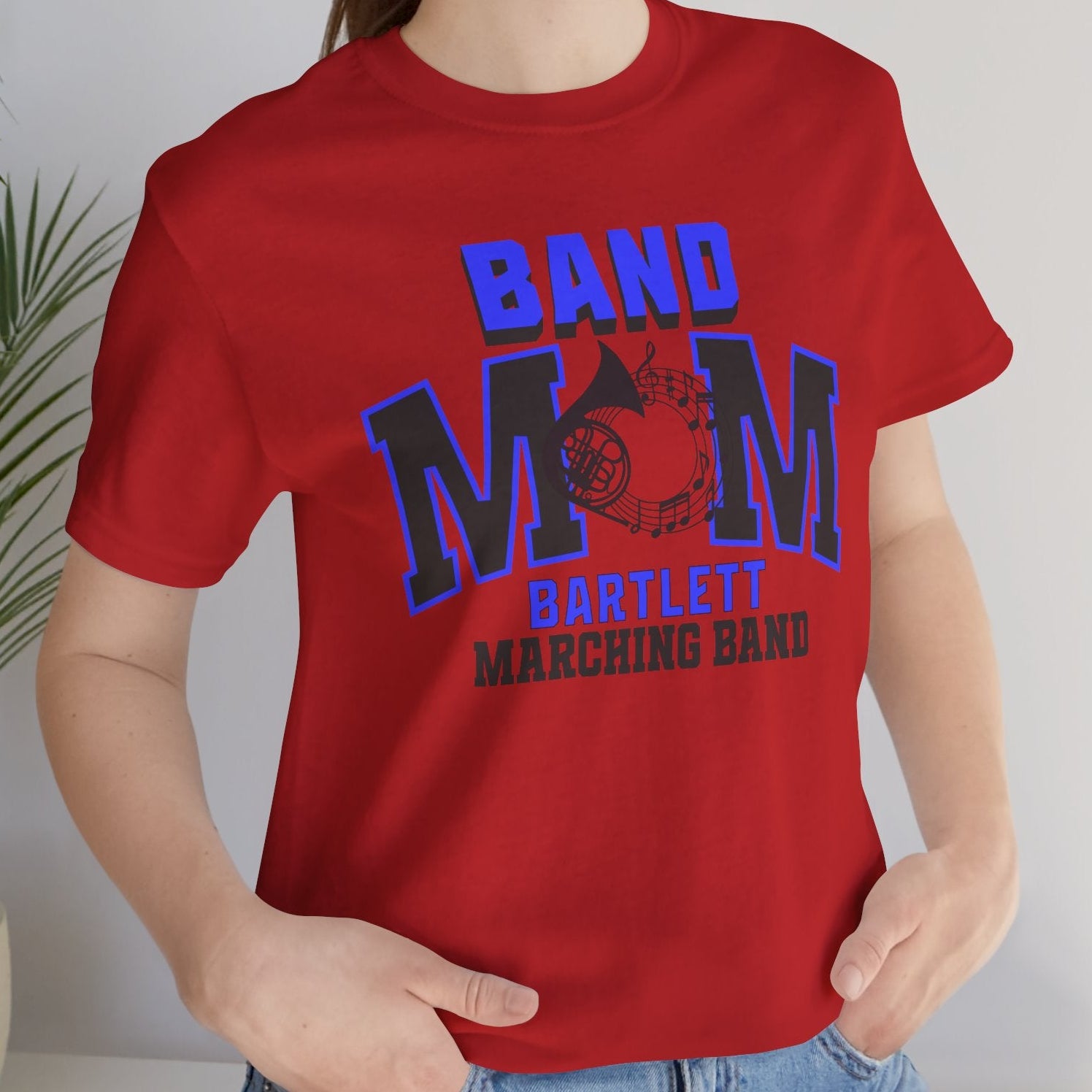 Person wearing a red 'Band Mom' t-shirt with text and graphics, standing indoors next to a plant.