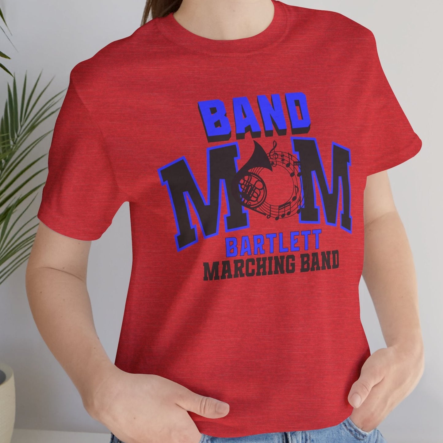 Person wearing a red 'Band Mom' t-shirt with blue and black text, standing indoors next to a plant.