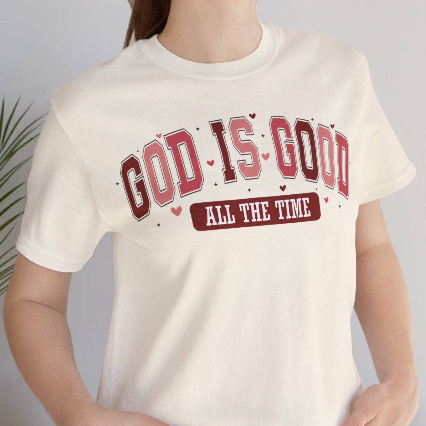 A woman smiling, wearing a white crew neck t-shirt with the text 'GOD IS GOOD ALL THE TIME' printed in red and brown colors.